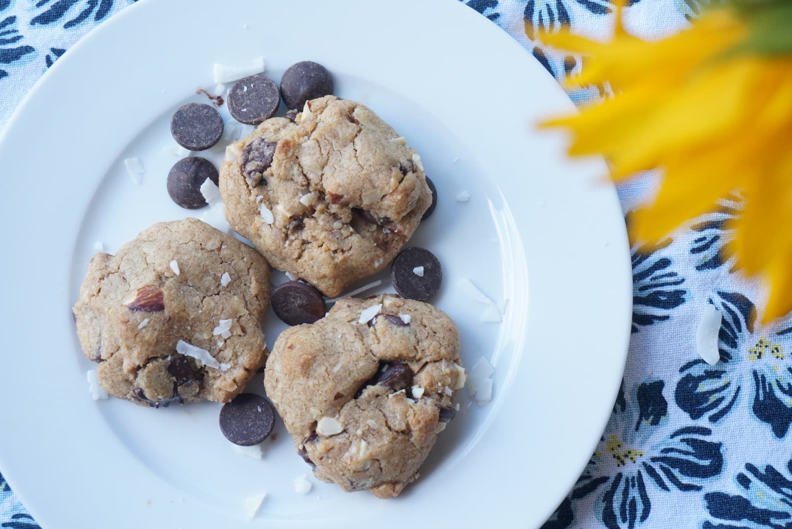 Three chocolate-chip and coconut cookies on a white plate beside yellow sunflowers.