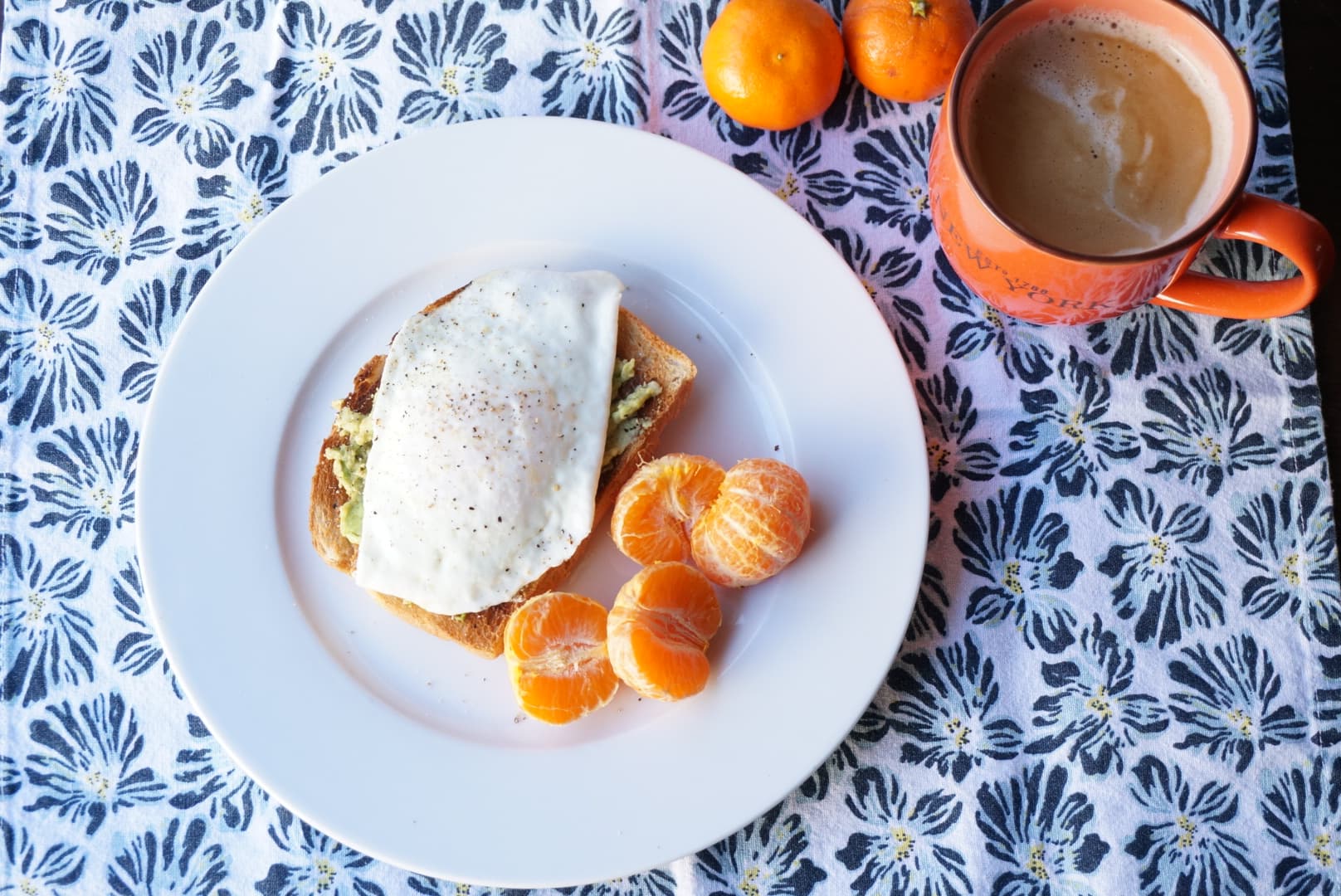 An egg over easy on avocado toast beside clementine slices and a mug of coffee, on a blue floral tablecloth.